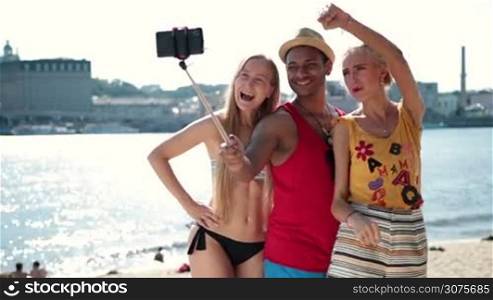 Happy friends making selfie photo on a selfie stick on the beach during summer vacation. Group of smiling friends posing and making selfie on smartphone