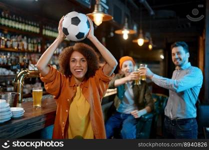 Happy friends rest at sports bar and drinking beer. Focus on overjoyed young woman holding soccer ball over head ready to throw into camera. Happy friends rest at sports bar, focus on woman holding soccer ball