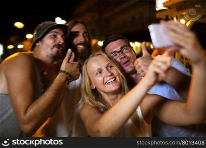 Happy friends selfie with thumbs up. Cheerful young people taking happy selfie with a mobile phone during night party. Jolly photo with thumbs up