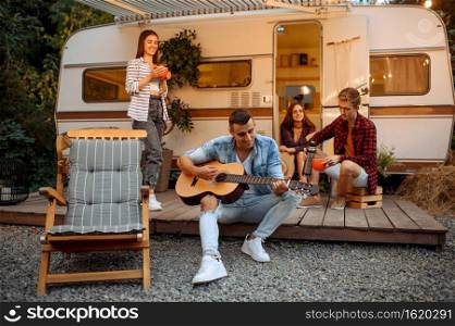 Happy friends sing songs with guitar on picnic at camping in the forest. Youth having summer adventure on rv, camping-car on background. Two couples leisures, travelling with trailer. Friends sing songs with guitar, picnic at camping