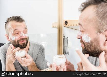 Happy funny adult guy applying moisturizer cream for male skin care. Man in front of bathroom mirror with anty aging lotion on face.. Man applying moisturizer cream in bathroom