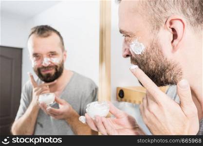 Happy funny adult guy applying moisturizer cream for male skin care. Man in front of bathroom mirror with anty aging lotion on face.. Man applying moisturizer cream in bathroom