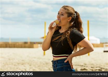 Happy funny young woman with long brown hair eating ice cream having fun enjoying her dessert during beautiful summer weather on beach. Young woman eating ice cream
