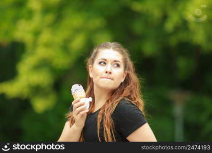 Happy funny young woman with long brown hair eating ice cream having fun enjoying her dessert during beautiful summer weather against green park. Young woman eating ice cream