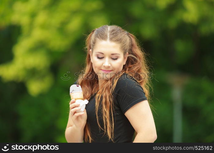 Happy funny young woman with long brown hair eating ice cream having fun enjoying her dessert during beautiful summer weather against green park. Young woman eating ice cream