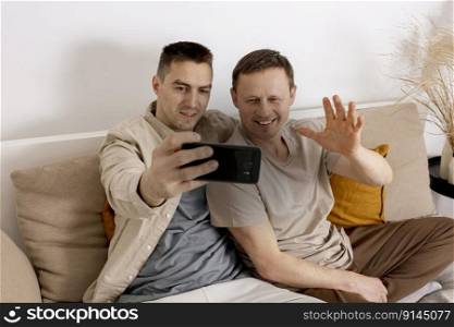 Happy gay couple with casual clothes holding smartphone and talking with friends online, having video call. Homosexual relationships and alternative love. Happy gay couple with casual clothes holding smartphone and talking with friends online, having video call. Homosexual relationships and alternative love.