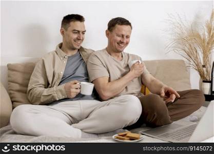 Happy gay couple with casual clothes spending time together at home, watching movie on the laptop and drinking tea. Two caucasian men relaxing. Homosexual relationships and alternative love. Happy gay couple with casual clothes spending time together at home, watching movie on the laptop and drinking tea. Two caucasian men relaxing. Homosexual relationships and alternative love.