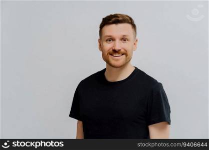 Happy ginger man in black t-shirt, pleasant expression. Studio shot, positive emotions. Grey wall backdrop.