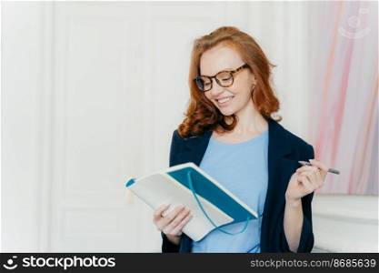 Happy ginger young female plans working schedule, writes in notebook, makes notes of useful information, holds pen, has smile on face, dressed elegantly, stands indoor, writes down her thoughts