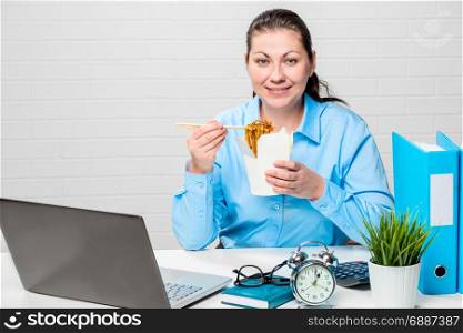 Happy girl accountant with a box of noodles and chopsticks at a table in the office
