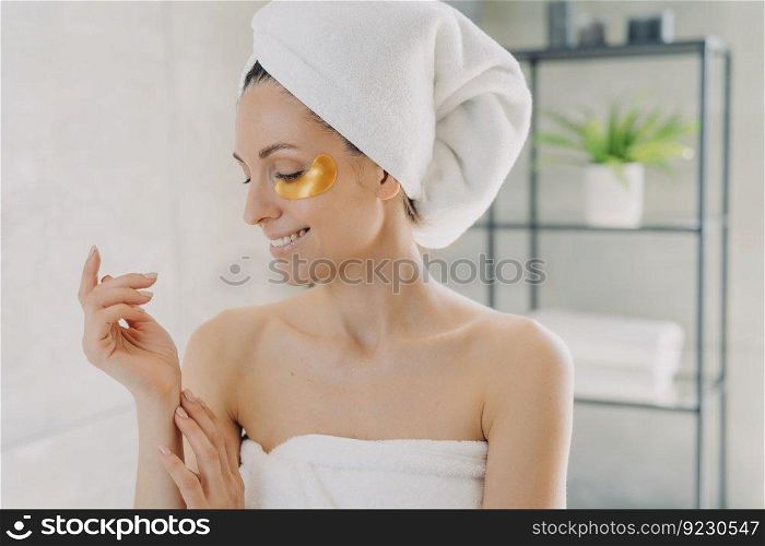 Happy girl applies golden eye patches and touches silky skin of her hands. Attractive european woman wrapped in towel after spa procedures. Young hispanic lady does hair and body care at home.. Girl applies eye patches and touches silky skin of her hands. Young lady does hair and body care.