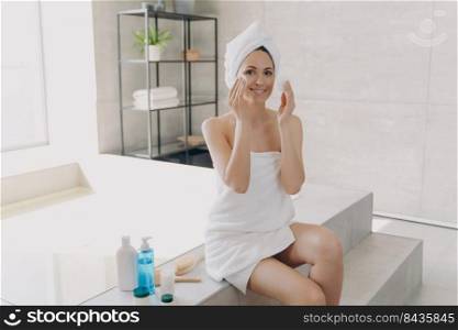Happy girl applying face lotion with cotton disks. Gorgeous caucasian woman wrapped in towel after bathing is sitting on bathtub. Young woman takes shower at home. Skin purifying and spa procedures.. Happy girl applying face lotion with cotton disks. Skin purifying and spa procedures at home.