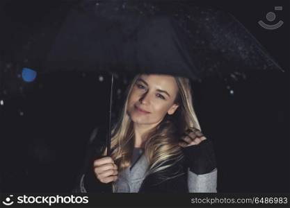 Happy girl at rainy night outdoors, portrait of a beautiful blond female standing with umbrella under rainfall, good mood in cold wet weather. Happy girl at rainy night outdoors