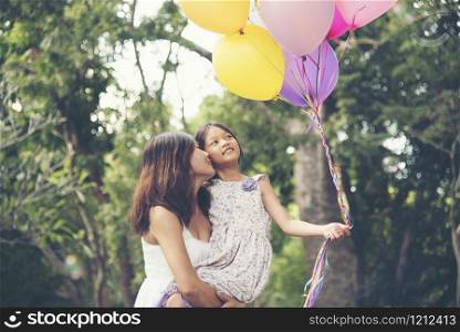 Happy girl holding colorful of air balloons on a green meadow with cloudy and blue sky. Mother hold daughter.