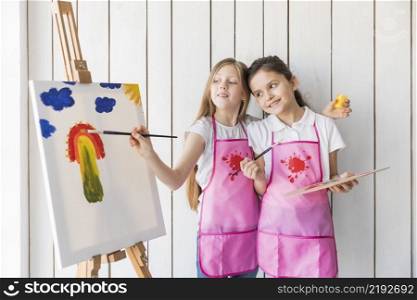 happy girl holding palette hand looking her friend painting canvas with brush