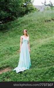 Happy girl in a turquoise long dress in a green park on a background of herbs, trees and rose bushes