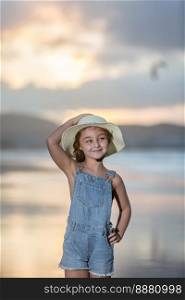 Happy girl in denim overall shorts touching hat and holding hand on waist while standing on Famara Beach near sea at sunset and looking away with smile in weekend in Lanzarote, Spain. Girl touching hat on beach