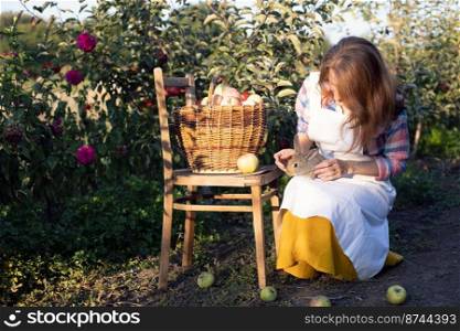 happy girl in the garden holds a rabbit in her arms and a basket of apples nearby. aesthetics of rural life 