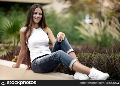 Happy girl in the park, cute young woman with beautiful long hair sitting on the parapet in the garden, conceptual photo of freshness of youth like a freshness of springtime nature 
