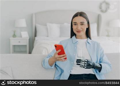 Happy girl is holding glass of water with cyber hand prosthesis. European woman with artificial arm is using smartphone at home. Healthy lifestyle after&utation. Futuristic medical technology.. Happy girl is holding glass of water with cyber hand prosthesis. Futuristic medical technology.