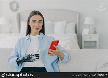 Happy girl is holding glass of water with cyber hand prosthesis. European woman with artificial arm is using smartphone at home. Healthy lifestyle after&utation. Futuristic medical technology.. Happy girl is holding glass of water with cyber hand prosthesis. Futuristic medical technology.