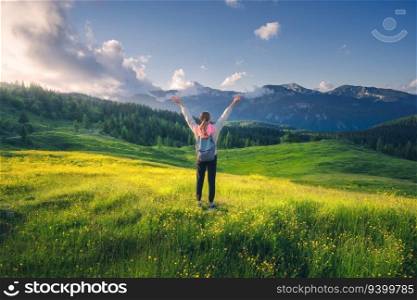 Happy girl on the hill with yellow flowers and green grass in beautiful alpine mountain valley at sunset in summer. Landscape with young woman with raised up arms in alps, trees, sky with clouds