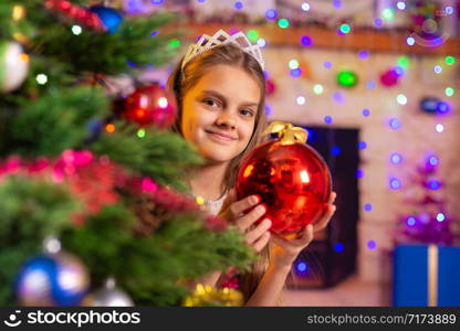 Happy girl peeps out from behind a beautiful Christmas tree with a ball in her hands