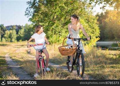 Happy girl riding on bicycle with her mother in meadow at sunset