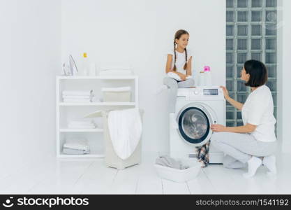 Happy girl sits at washing machine, has pleasant conversation with mother, being in laundry room, woman loads washer with dirty clothes, white shelf near with neatly folded linen, iron, detergents