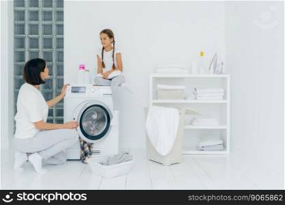Happy girl sits at washing machine, has pleasant conversation with mother, being in laundry room, woman loads washer with dirty clothes, white shelf near with neatly folded linen, iron, detergents