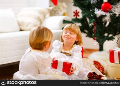 Happy girl sitting near Christmas tree and presenting gift to her sister