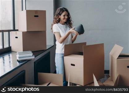 Happy girl unpacking cardboard boxes and taking the vase. Woman in casual outfit. Smiling attractive european woman packing boxes to move. Relocation concept.. Happy girl unpacking cardboard boxes and taking the vase. European woman packing boxes to move.