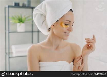 Happy girl uses gold eye patches, cares for silky hands. Attractive European in towel post-spa. Young Hispanic lady does hair and body care at home.