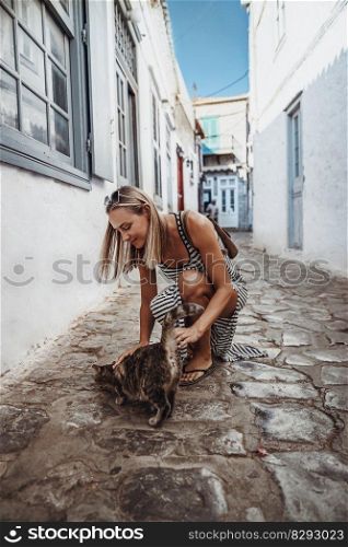 Happy girl walking on a cute white narrow streets of Greece. Petting nice cat. Enjoying active holidays in Europe.. Pretty woman spending vacation in Greece