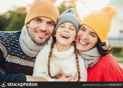 Happy girl with pigtails, wears warm knitted sweater, stands between father and mother, laugh happilly, have sincere smiles on their faces. Relaxed family have holidays, spend nice time together