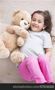 Happy girl with teddy bear lying on wooden floor at home