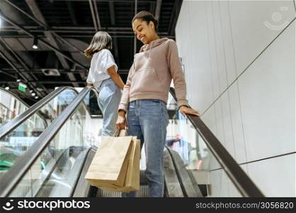 Happy girls with purchases in bags on escalator, clothing store. Women shopping in fashion boutique, shopaholics, shoppers in sale center. Happy girls with purchases in bags on escalator