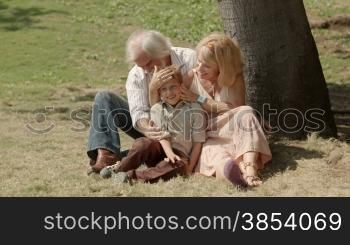 Happy grandfather and grandmother with grandson sitting on grass under tree in park