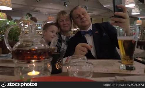 Happy grandma, grandpa and grandson taking family selfie with cellphone during celebratory dinner in the restaurant