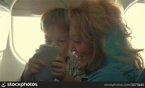 Happy grandmother holding little grandson on the lap and watching how using smart phone. Family travel by plane