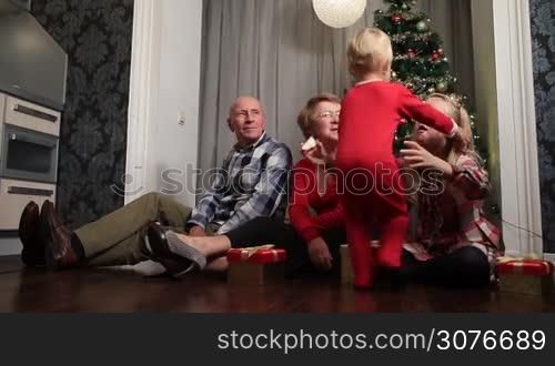 Happy grandparents and little girl sitting on the floor with gift boxes in front of Christmas tree at home. Cute little toddler boy running to his big sister and embracing her. Smiling family reunion celebrating winter holidays together.