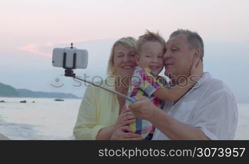 Happy grandparents and little grandson taking mobile selfie at the seaside. Grandpa holding boy and they using selfie stick to make great a picture