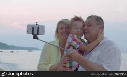 Happy grandparents and little grandson taking mobile selfie at the seaside. Grandpa holding boy and they using selfie stick to make great a picture