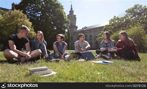 Happy group of college students communicating and sharing ideas with each other on campus lawn while relaxing after studies finished. Positive university friends chatting about latest news and gossips after class as they sit on grass in the park