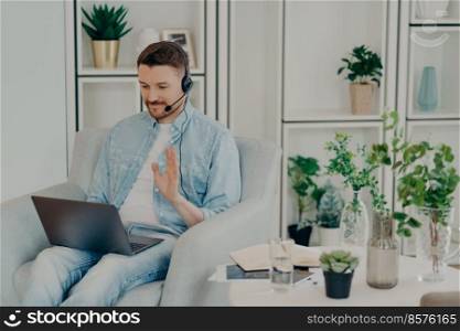 Happy guy in headset waving hand at laptop webcam while sitting in chair at home and chatting online with friend, smiling male freelancer having video call with colleagues while working remotely. Caucasian young man relaxing at home and chatting online, waving hand