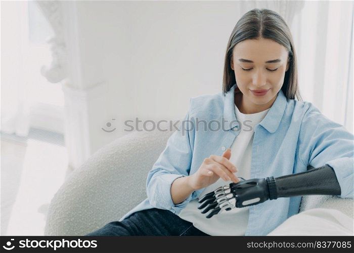 Happy handicapped girl is setting up her artificial limb and pressing buttons. Young european woman with cyber hand at home. Modern robotic bionic prosthesis and orthopedic technology. Copy space.. Happy handicapped girl is setting up her artificial limb and pressing buttons. Copy space.