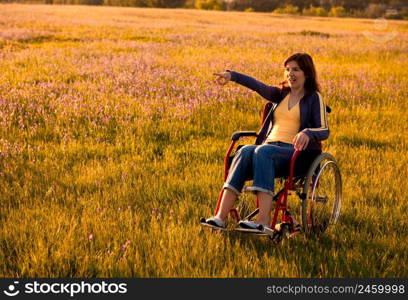 Happy handicapped woman on a wheelchair over a green meadow pointing and looking away