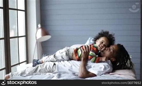 Happy handsome african american father with dreadlocks lying on bed and playing with cheerful toddler son. You are my world! Side view of playful young african man playing with his little boy and smiling while lying on his back on the bed.