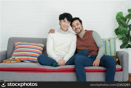 Happy handsome Asian young gay couple wearing sweater embracing his boyfriend looking at camera and sitting on sofa together at living room at home. Romance Lgbt family in Valentine day