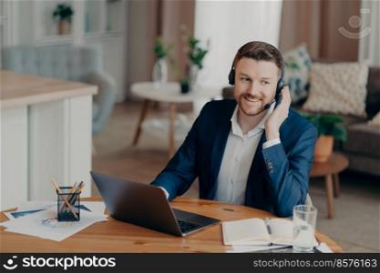 Happy handsome man business professional sitting in living room at his cozy workplace while enjoying web conference or online meeting, using laptop and headset. Remote work and freelance concept. Smiling handsome young businessman in headset studying online at home